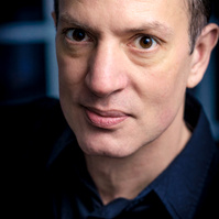 Close-up professional headshot of a man in a dark shirt with an intense expression, set against a softly lit, dark background. Perfect for actor profiles or creative branding.