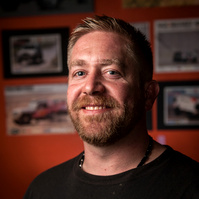 Portrait of a smiling man in a racing suit, set against a blurred background featuring motorsport imagery. Perfect for motorsport profiles, racing promotions, or competitive branding.