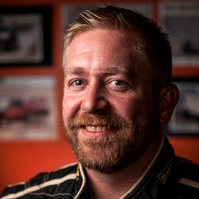 Portrait of a smiling man in a racing suit, set against a blurred background featuring motorsport imagery. Perfect for motorsport profiles, racing promotions, or competitive branding.