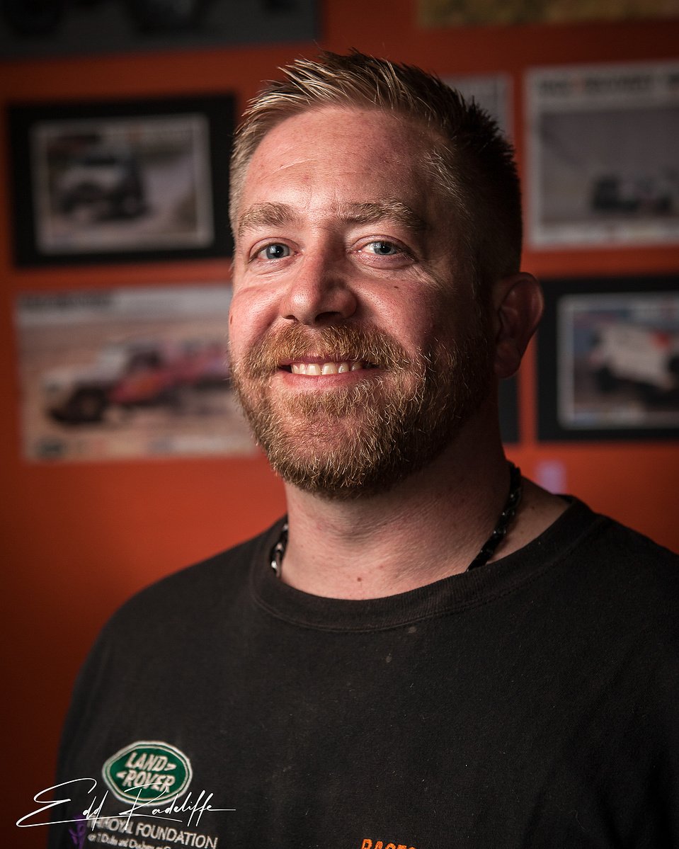 Portrait of a smiling man in a racing suit, set against a blurred background featuring motorsport imagery. Perfect for motorsport profiles, racing promotions, or competitive branding.