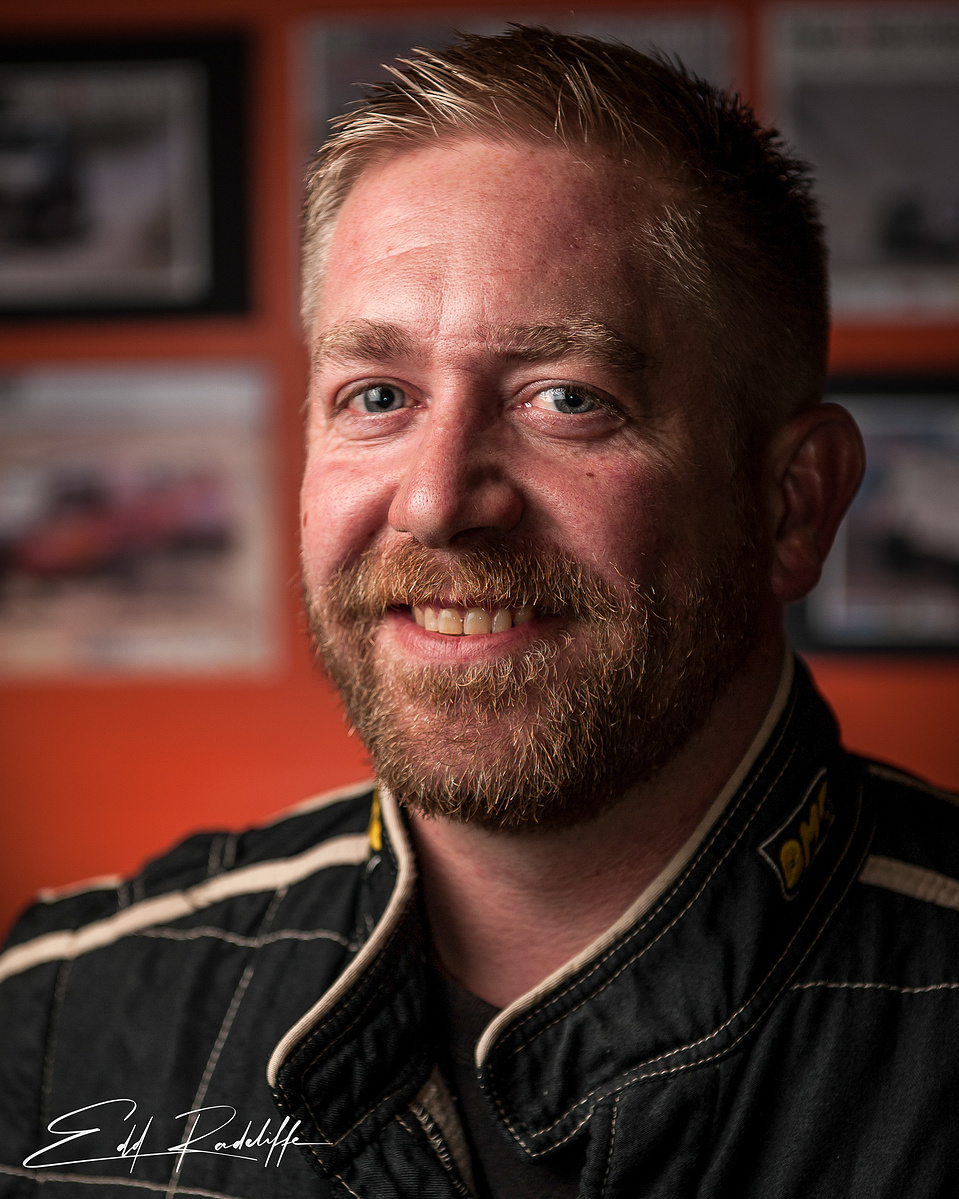 Portrait of a smiling man in a racing suit, set against a blurred background featuring motorsport imagery. Perfect for motorsport profiles, racing promotions, or competitive branding.