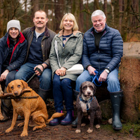 Group of four adults seated outdoors on a wooden bench with three dogs in a forest during autumn. Casual attire, natural setting, and relaxed atmosphere with two retrievers and one mixed-breed dog.