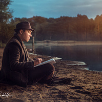Atmospheric portrait of a man in a coat and hat sitting by a tranquil lake at sunrise, sketching in a notebook. Ideal for creative storytelling, artistic branding, or contemplative themes.