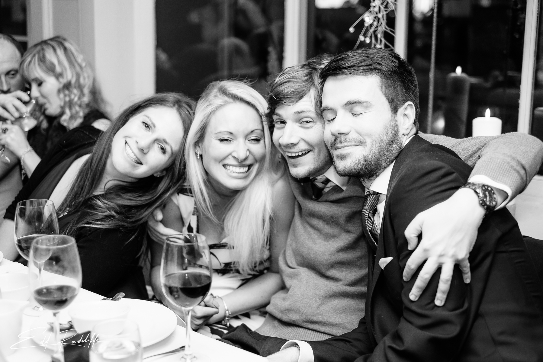 Friends embrace while sitting at wedding reception meal, smiling at camera at Chiswell Street Dining Rooms, Islington for Edd Radcliffe Photography - documentary style wedding photography
