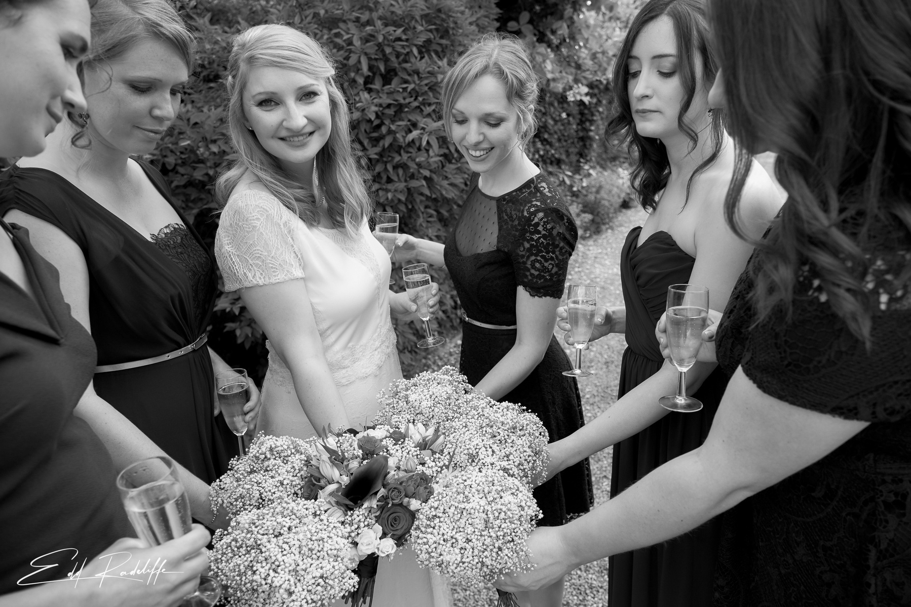 Bride smiles at camera while her and her bridesmaids stand in a circle holding their bouquets together at the Reid Rooms, Chelmsford for Edd Radcliffe Photography - documentary style wedding photography