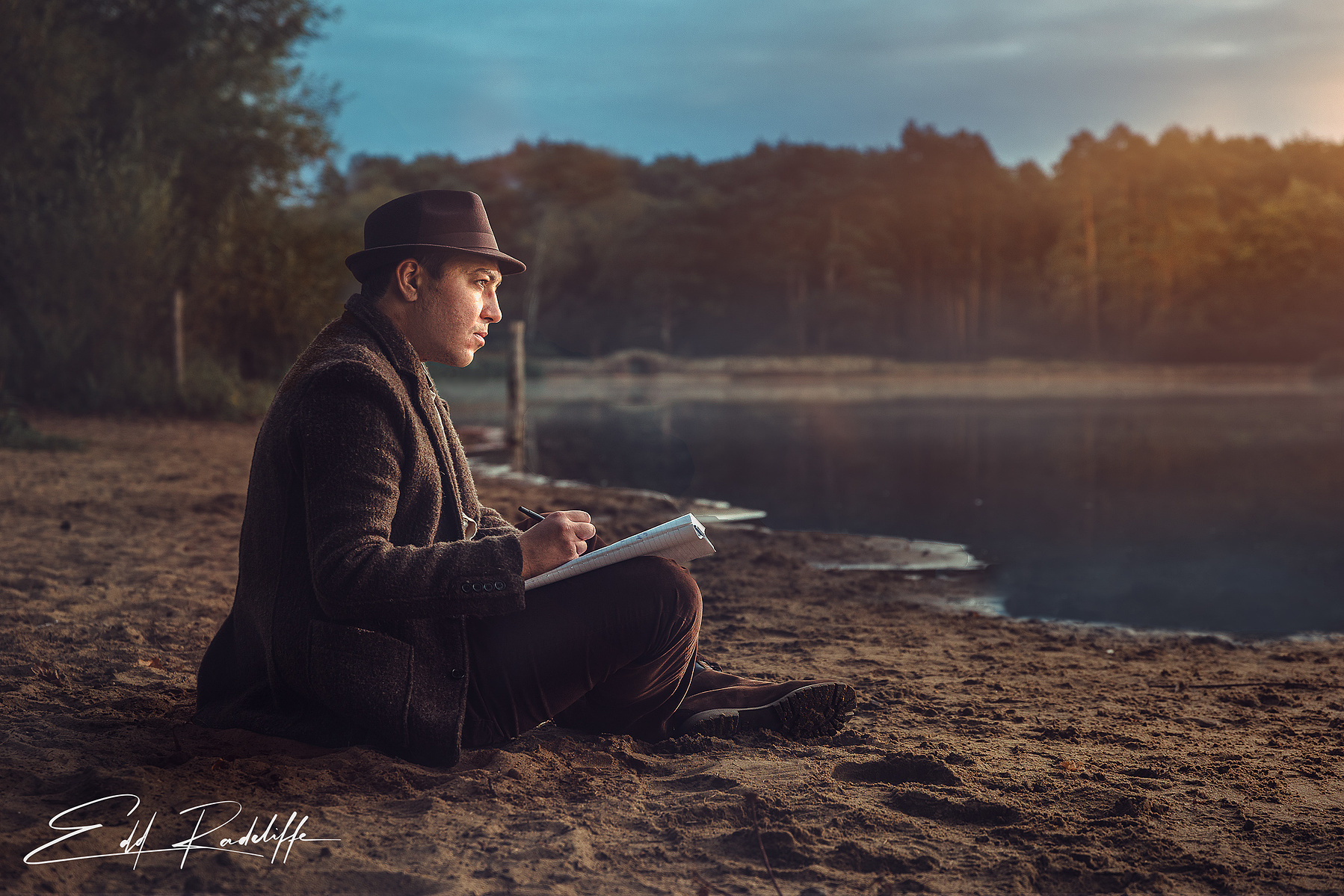 Atmospheric portrait of a man in a coat and hat sitting by a tranquil lake at sunrise, sketching in a notebook. Ideal for creative storytelling, artistic branding, or contemplative themes.