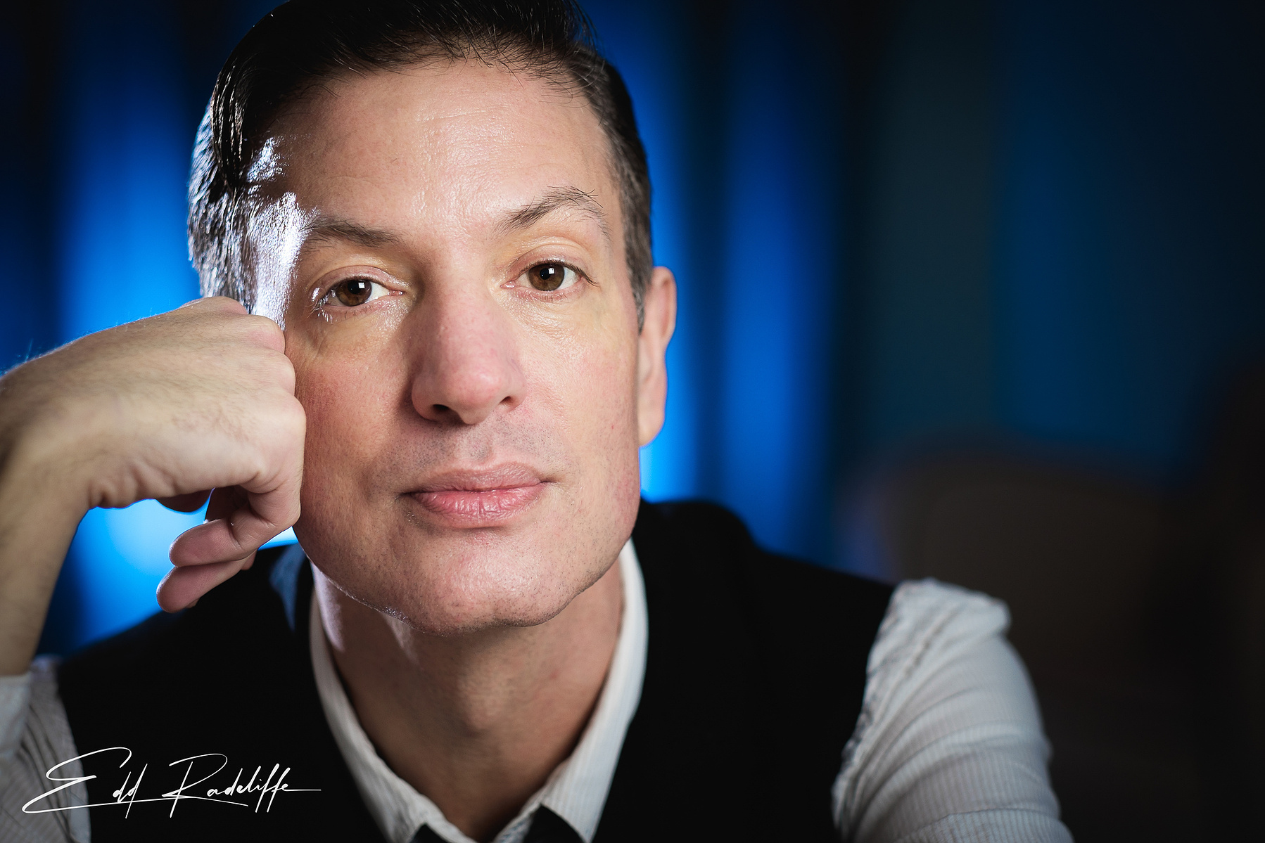 Close-up portrait of a thoughtful man resting his chin on his hand, wearing a vest and tie, with a blue-lit background. Perfect for creative profiles, artistic branding, or professional use.