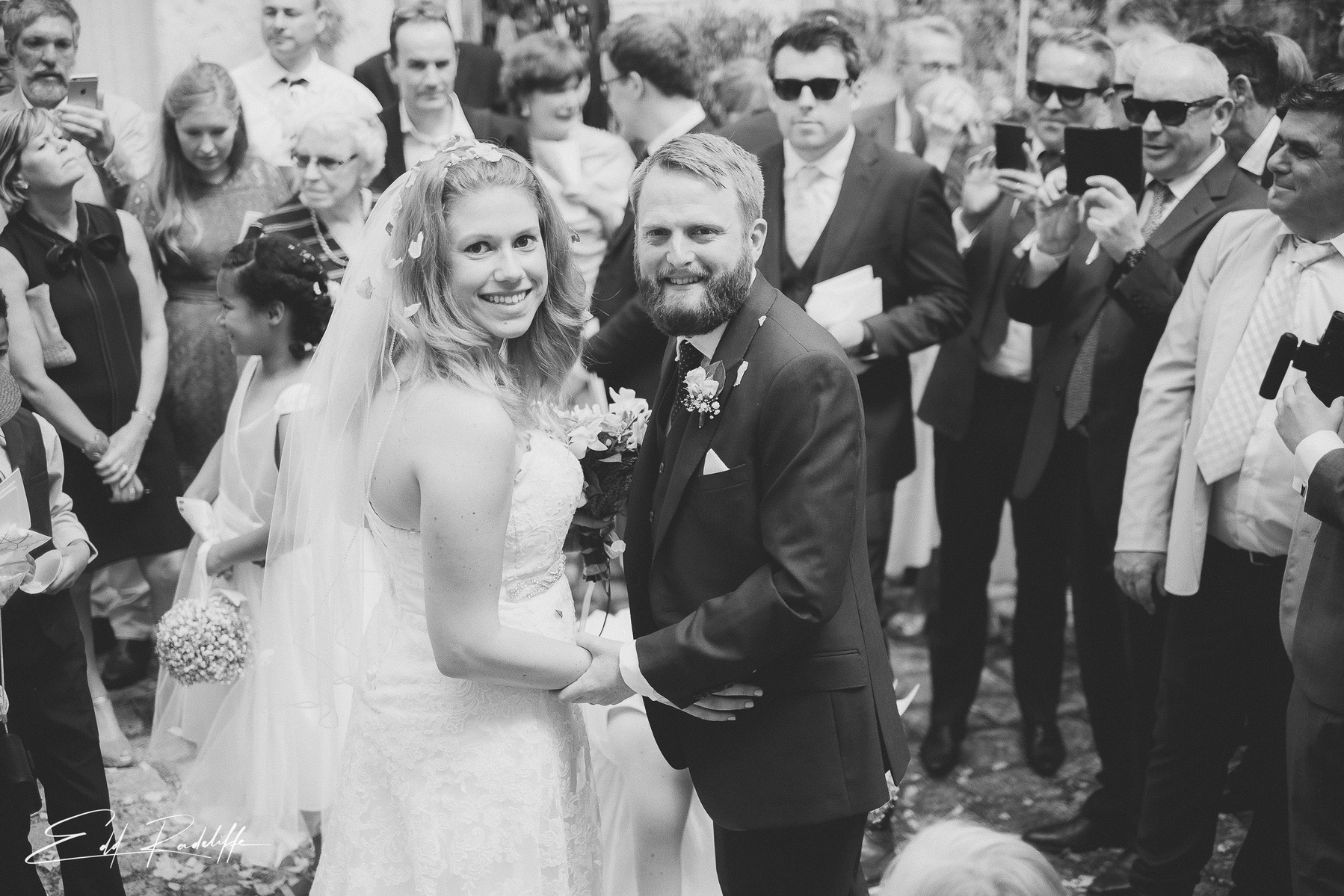 Bride and Groom look back at camera surrounded by friends and family taking photos at Hôtel de Cours de Thomazeau in Castilliones for Edd Radcliffe Photography - documentary style wedding photography