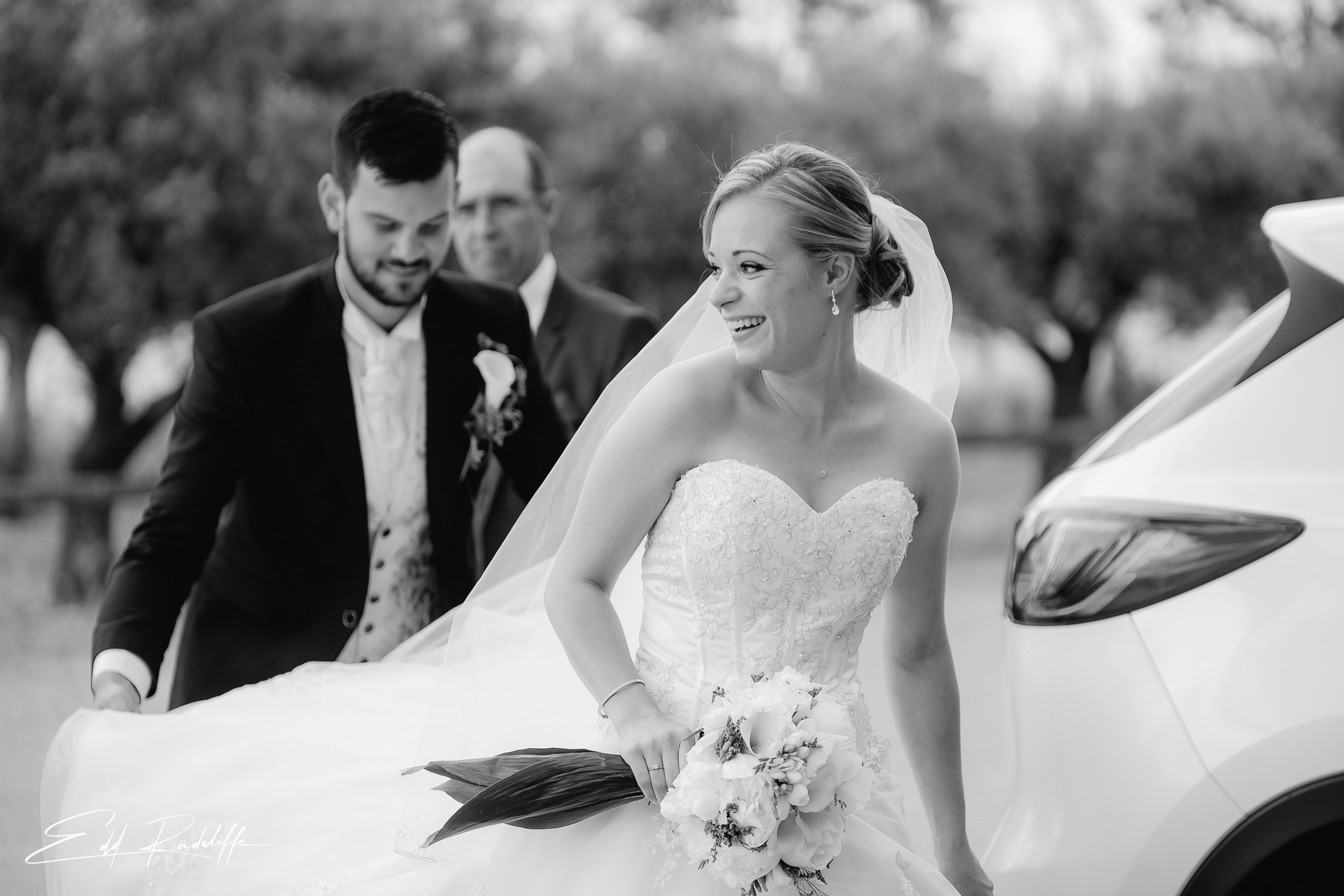 Groom holds train of Brides dress as she smiles at Garons, France for Edd Radcliffe Photography - documentary style wedding photography
