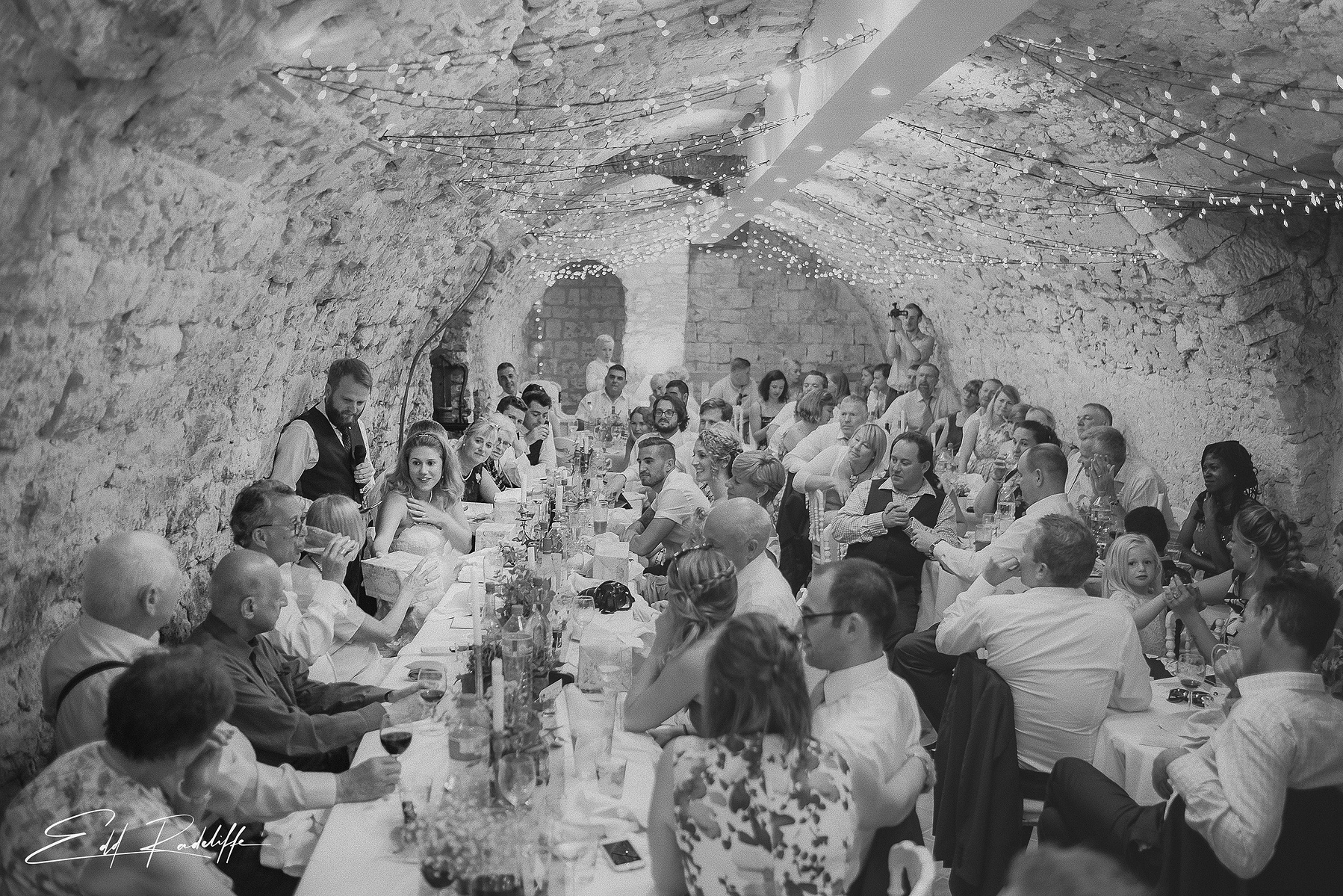 Bride hands gift to wedding guest while Groom looks on during wedding breakfast in beautiful underground vault at Hôtel de Cours de Thomazeau in Castilliones for Edd Radcliffe Photography - documentary style wedding photography