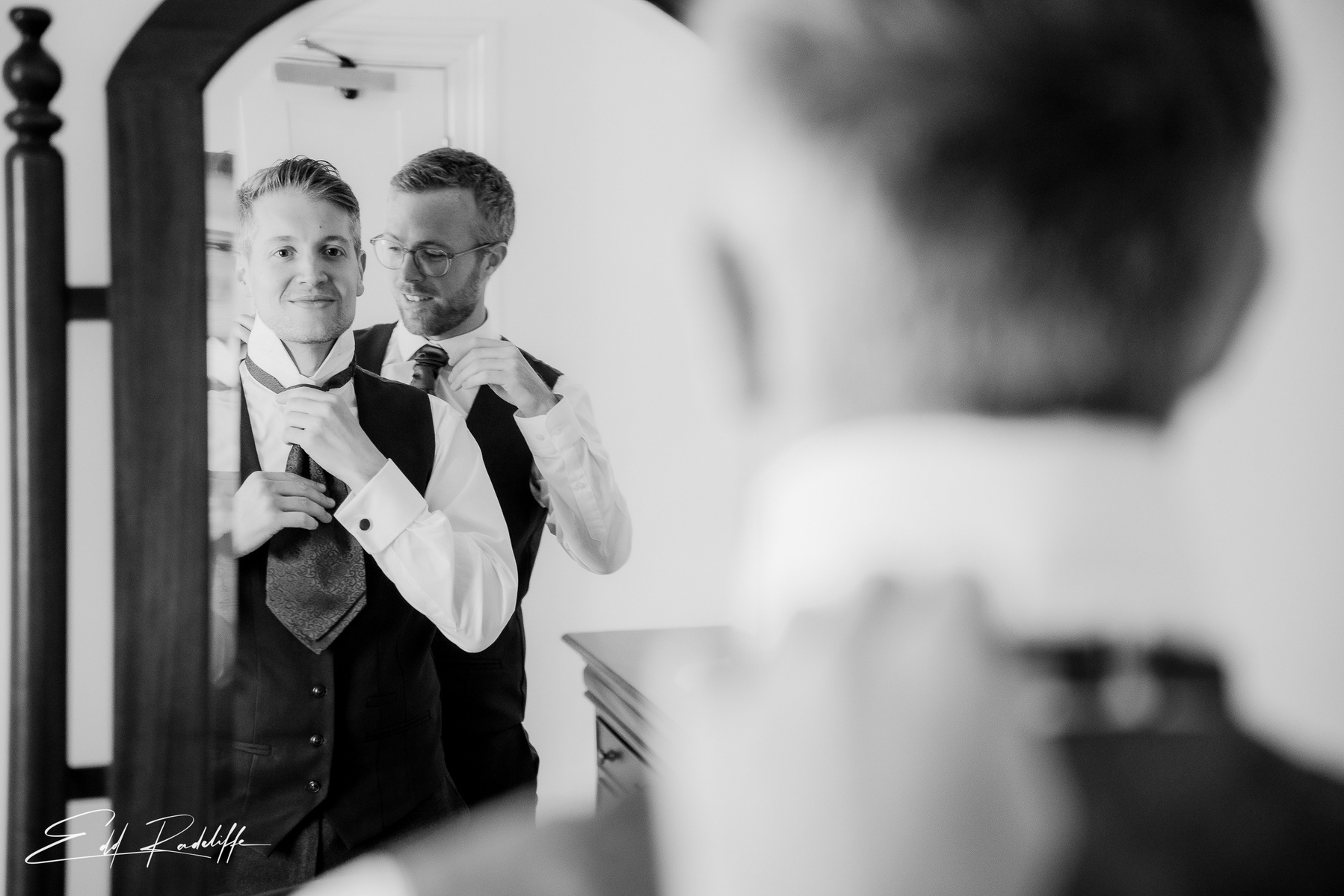 Groom looks at his reflection in mirror while doing up his tie at Groomes in Hampshire for Edd Radcliffe Photography - documentary style wedding photography