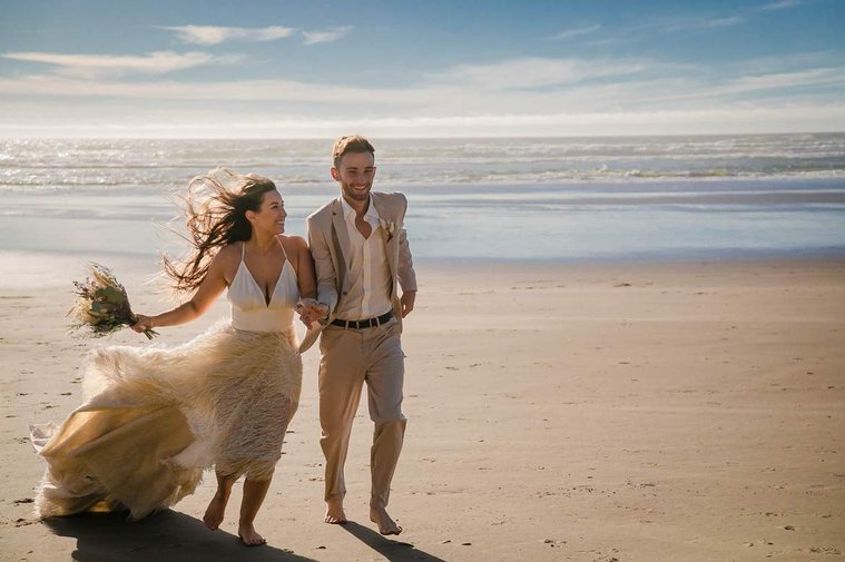 Couple in wedding attire run along a beach at the Oregon Coast.