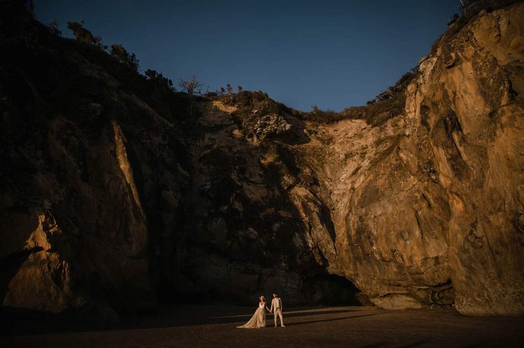 Couple stands holding hands in front of the sunlit rocks at Hug Point Beach, Oregon.