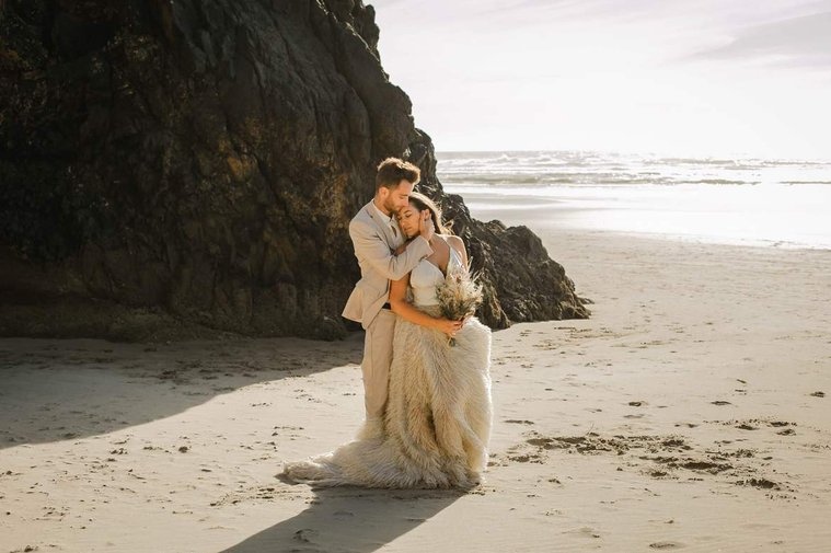 Bride and groom in summer wedding outfits  hold each other close on a beautiful Oregon Coast beach.