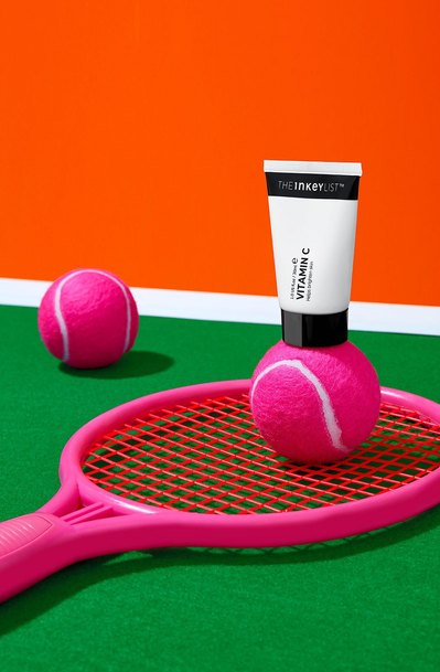 a beauty product delicately balanced on a pink tennis ball, itself resting on a pink rack within a tennis court. This visually harmonious composition is skillfully captured by David Lineton, an expert in London makeup still life photographer