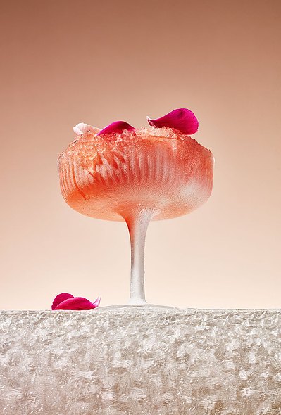 A refreshing composition featuring a drink glass completely filled with ice pieces, adorned with a flower leaf on top, and an additional flower leaf placed just outside the glass. This visually appealing scene is skillfully photographed by David Lineton. 