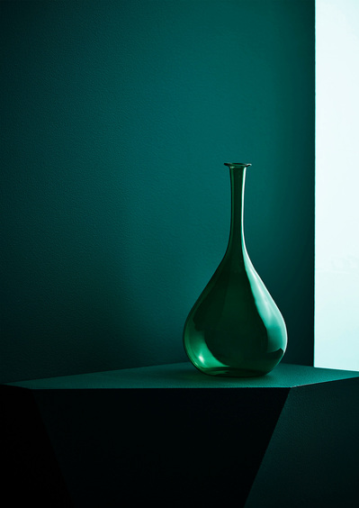 A captivating composition featuring a greenish, uniquely shaped bottle placed on a greenish surface with a matching background. This visually appealing image is skillfully captured by David Lineton, an expert creative product photographer in London