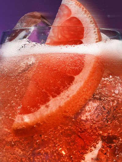 A vibrant composition capturing a reddish-colored drink glass, with the top half elegantly showcased, featuring a visible lemon piece and ice on top. This visually appealing image is skillfully photographed by David Lineton, an expert in drinks photograph