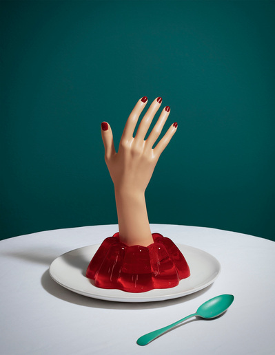 A delectable scene featuring a red jelly cake on a plate, adorned with a hand cutout displaying five fingers. This visually enticing image is captured by David Lineton, a specialist in modern still life photography in London