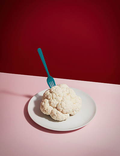 A tastefully arranged composition featuring cauliflower placed in a white plate with a fork, set on a table against a striking red background. This visually appealing image is skillfully captured by David Lineton, a modern still life photographer in Londo