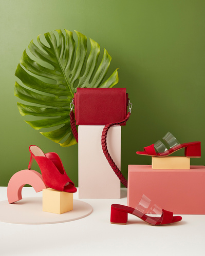 A dynamic composition featuring three red shoes artfully arranged on an object, with a red bag on top, set against a solid surface and complemented by a green background. Skillfully captured by David Lineton, an expert in accessories photography in London
