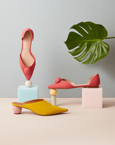 A balanced composition showcasing three shoes, two pink and one yellow, creatively arranged on surfaces. The scene is enhanced with the addition of a green leaf on top. Skillfully captured by David Lineton, an expert in accessories photography in London.