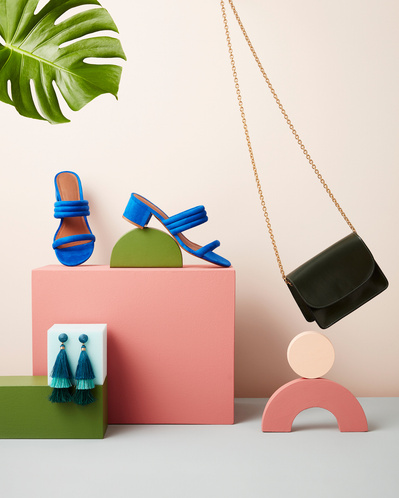 A stylish composition featuring a black purse elegantly hung with two blue small shoes placed on a surface. One shoe is positioned vertically, and the other is artfully placed on a small cup-shaped object. The scene is adorned with a green leaf on top. Sk