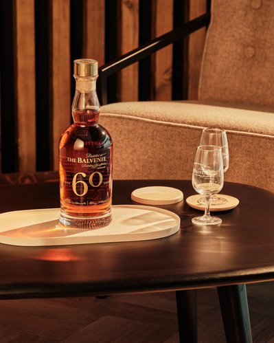The Balvenie 60 Years bottle is set in a lifestyle setting with two drams of whiskey from the bottle sitting on marble coasters next to the 60 YO Single Casked Whiskey. 