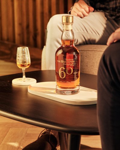 The Balvenie 60 Years bottle is set in a lifestyle setting with a drams of whiskey from the bottle sitting on marble coasters next to the 60 YO Single Casked Whiskey with people sitting and socialising over a dram 