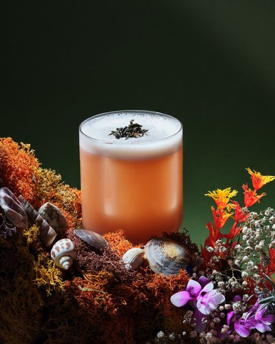 A delightful composition featuring a drink glass placed on a layer of flowers and leaves, with the top layer resembling foam. This visually engaging image is skillfully photographed by David Lineton, an expert in drinks photography in London. 