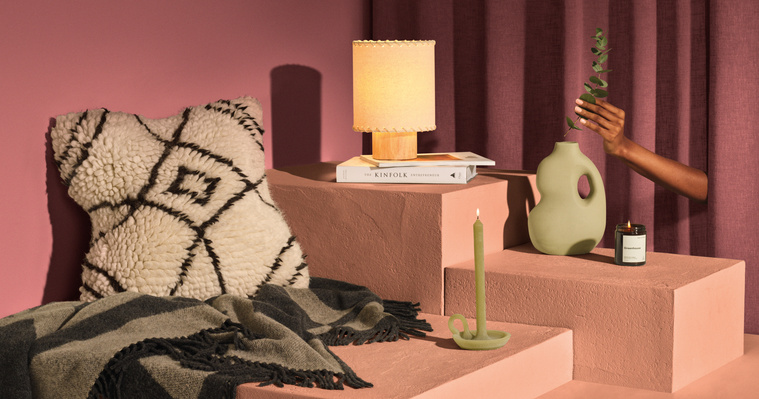 Gifting essentials from Made.com. A red set with peachy-toned plinths homes vases, cushions, throws candles and books with a hand placing a piece of eucalyptus in the Vase.