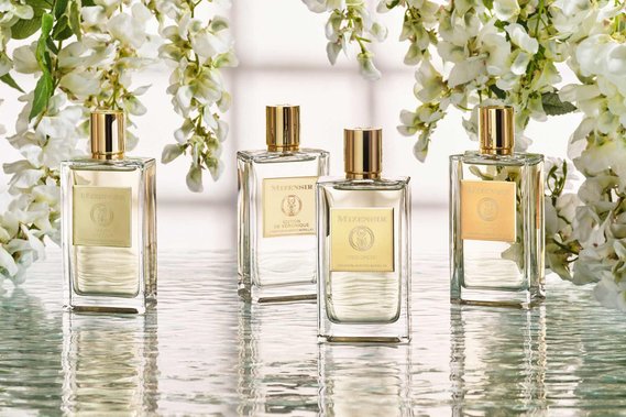 Mizensir Perfume bottles sit on water ripples as white wisteria flowers are dangling over the water ripples. Backlit by window light the bottles look as though they are floating in the water. 