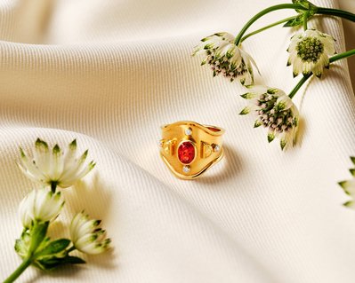 A luxurious composition featuring a gold ring adorned with red and white pearls, delicately placed in between flowers. This visually opulent arrangement is skillfully photographed by David Lineton, an expert in luxury product photography in London.