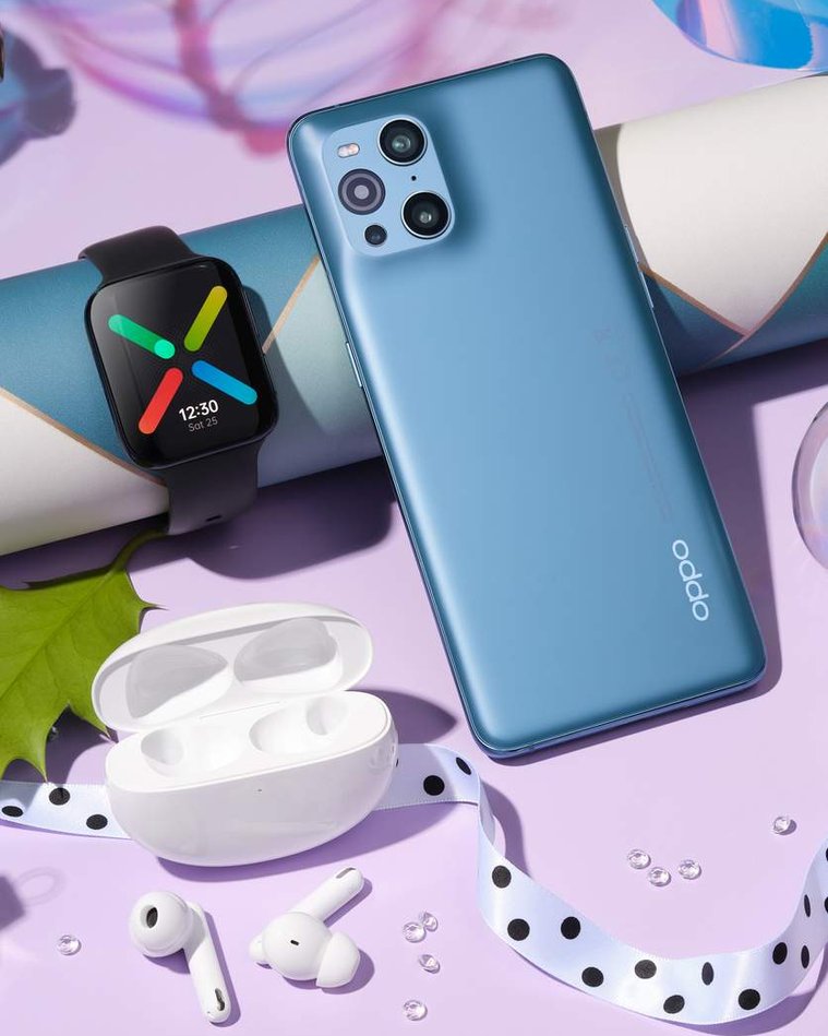 Oppo phone, digital watch face illuminated and earbuds photographed on the pink ultraviolet festive set with holly leaves and wrapping paper adding festive touches.