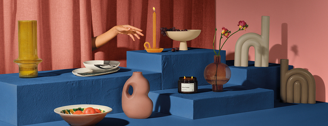 A vibrant, Red and Blue plinth set. Vases and small home accessories are placed on varying heights with a hand appearing from the Red fabric curtain reaching for a candle that is lit.