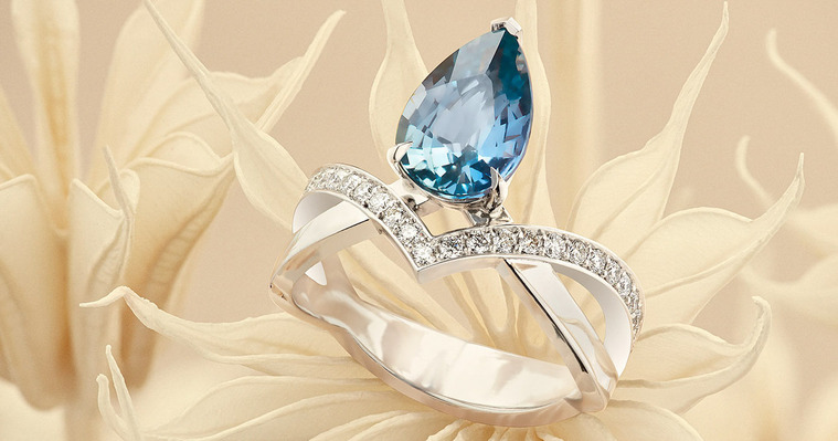A vibrant pear drop, teardrop-shaped gemstone sits within a fancy diamond setting. The ring is placed on dried flower stems. The soft peachy tones offset the polished platinum surfaces.