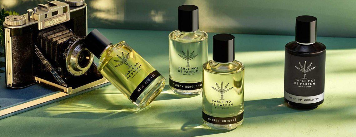 Sunset lighting casts light streaks and long refracted light shadows around the four Parle Moi De Parfum bottles. An old vintage camera props up a fragrance bottle. The green tones add a summer feel to the image 