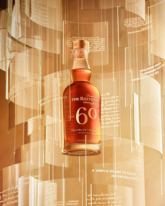The Hero key visuals of The Balvenie 60 Years single-casked Malt whiskey. Paines of glass with etching of quotes surround the bottle. A warm wash of honey glow lighting contrasts the tones and colour of the whiskey bottle.
