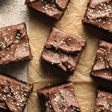 Brownies placed on a sheet of brown parchment paper and topped with chocolate frosting and sesame seeds.