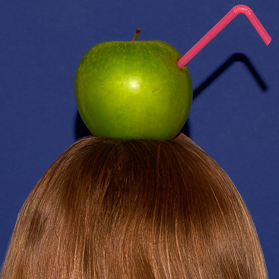 Detail of the back of the head of a woman with medium blond hair with a green apple and a pink straw on top, in front of a blue background
