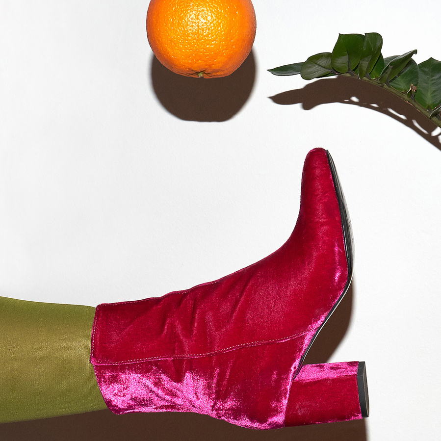 Detail of a pink woman's leg wearing a pink velvet ankle boot and green tights kicking an orange in the air in front of a white wall next to a plant