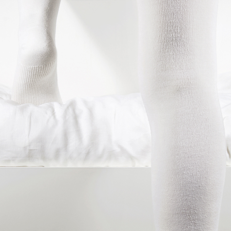 Close up of the backsides of a woman's legs wearing white tights stepping up a white drawer filled with white bedsheets