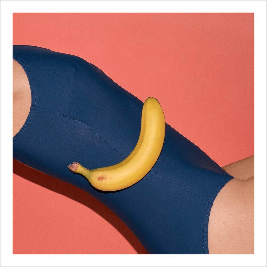 Part of a woman wearing a petrol colored bathing suit with a yellow banana in front and an apricot colored background