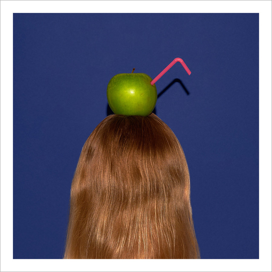 Back of the head of a woman with medium blond hair with a green apple and a pink straw on top, in front of a blue background
