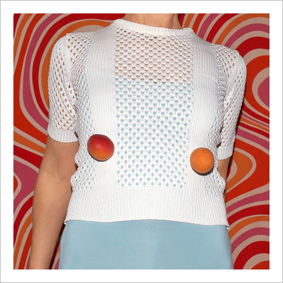 The upper body of a woman wearing a white knitted top with two apricots in front and a psychedelic retro orange pink pattern in the back