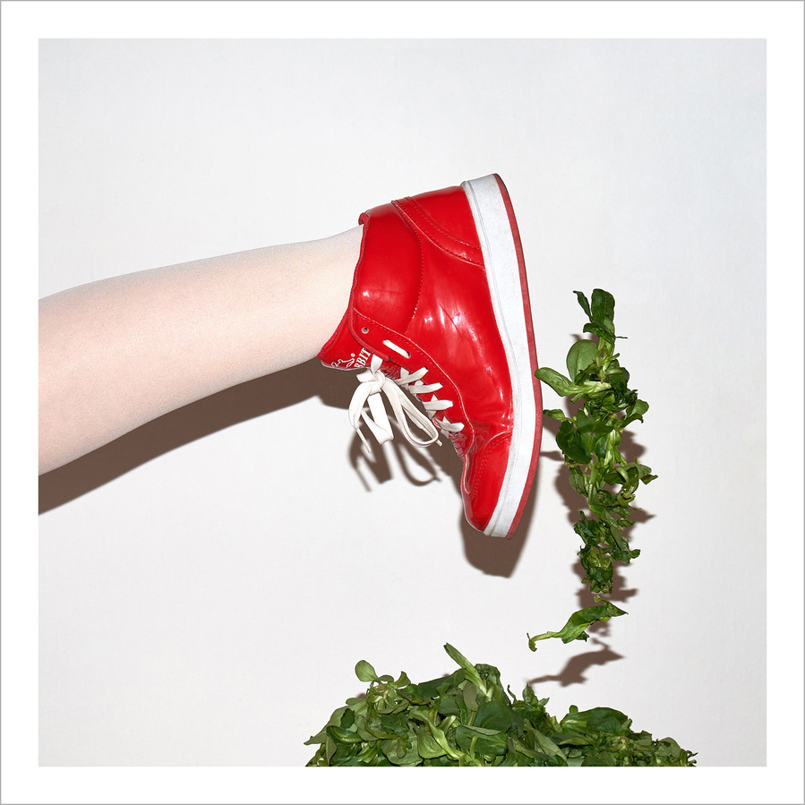 Leg of a woman wearing a bright red sneaker and white tights moving out of the image on heap of lamb's lettuce, green lettuce flying in the air in front of a white wall