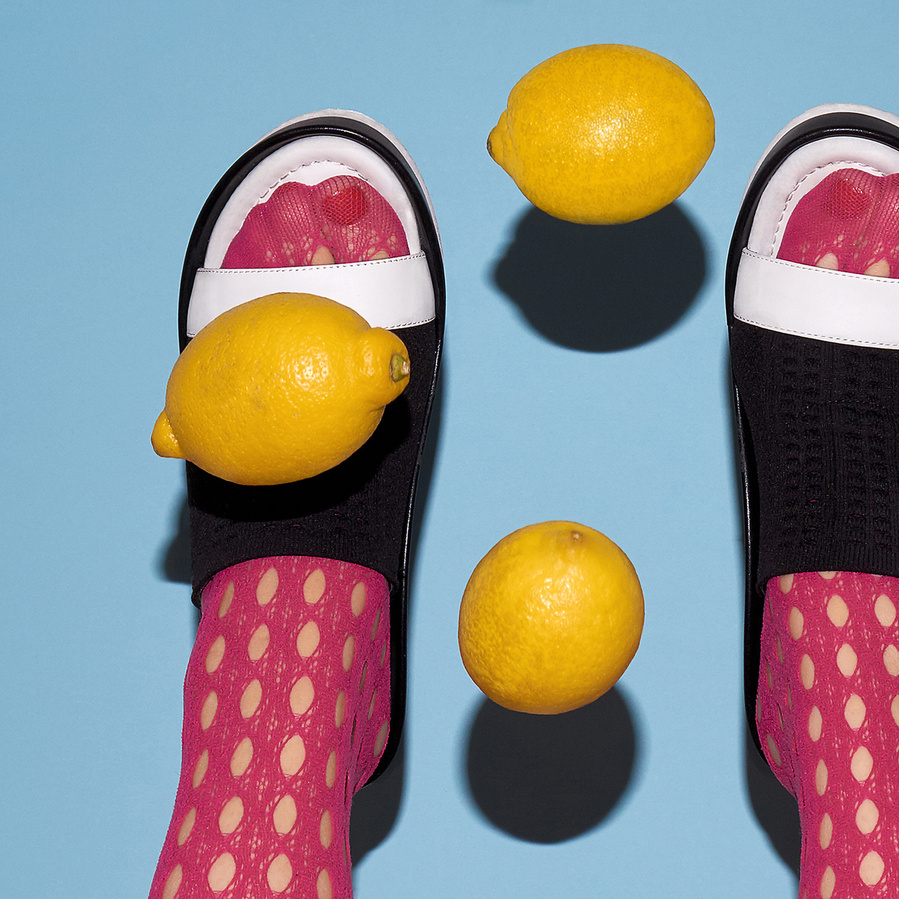 Detail of two female legs wearing pink tights and black and white shoes with three lemons flying in the air in front of a light blue background
