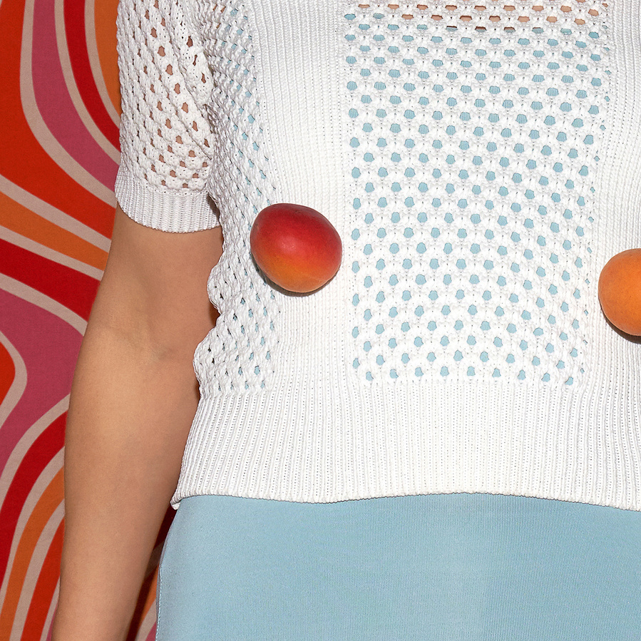 Detail of the upper body of a woman wearing a white knitted top with two apricots in front and a psychedelic retro orange pink pattern in the back