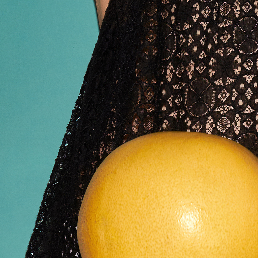 Detail of a black lace dress with a flying yellow pomelo in front and a petrol colored background