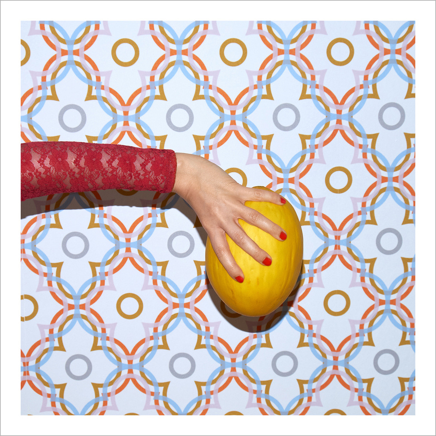 A female arm wearing a pink lace shirt, her hand with varnished red finger nails holding a yellow melon in front of a colorful patterned wall paper